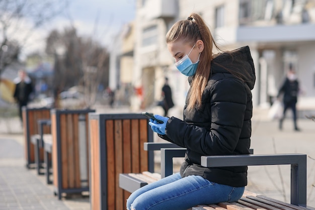 Joven mujer sentada en un banco de la calle con máscara médica y ropa negra