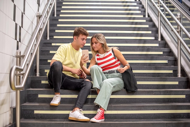 Joven y mujer sentada en las escaleras de una estación de metro Ella muestra algo en su teléfono celular