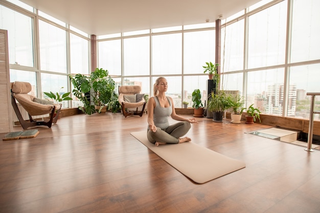 Joven mujer serena cruzando las piernas mientras está sentado en la estera en pose de loto y practicando ejercicios de meditación en el suelo en el entorno hogareño