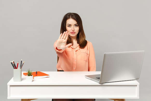 Joven mujer seria con ropa de color pastel que muestra un gesto de parada con la palma de la mano diciendo No hay trabajo en el escritorio blanco con una computadora portátil aislada en un fondo gris. Logro el concepto de carrera empresarial. Simulacros de espacio de copia.