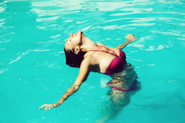 Joven mujer sexy o niña con cara bonita y cabello mojado nadando en la piscina con agua azul día soleado de verano al aire libre