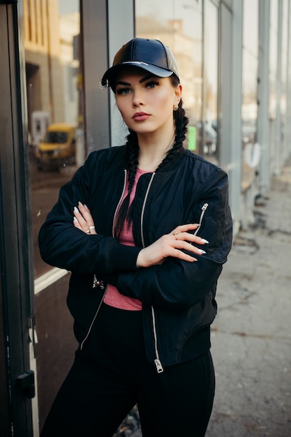 Joven mujer sexy con trenzas vestida con un pantalón negro y una chaqueta de bombardero negra, gorra de cuero.