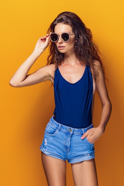 Joven mujer sexy vestida con traje de baño y pantalones cortos de jeans azul, gafas de sol posando en el amarillo.