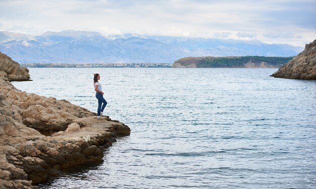 Joven mujer sola en la pedregosa orilla del mar, disfrutando de hermosas vistas