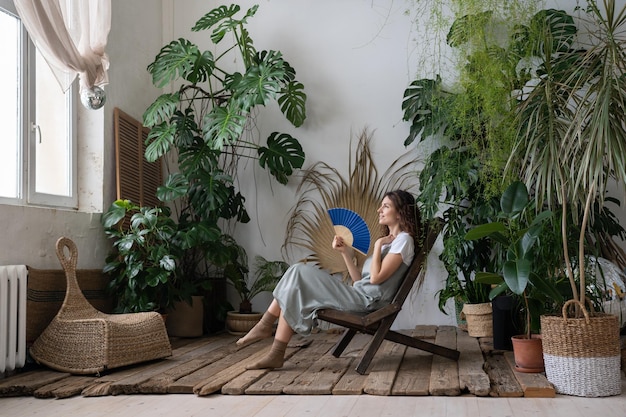 Joven mujer soñadora y relajada que usa un abanico de papel mientras se relaja en un hermoso y refrescante jardín casero