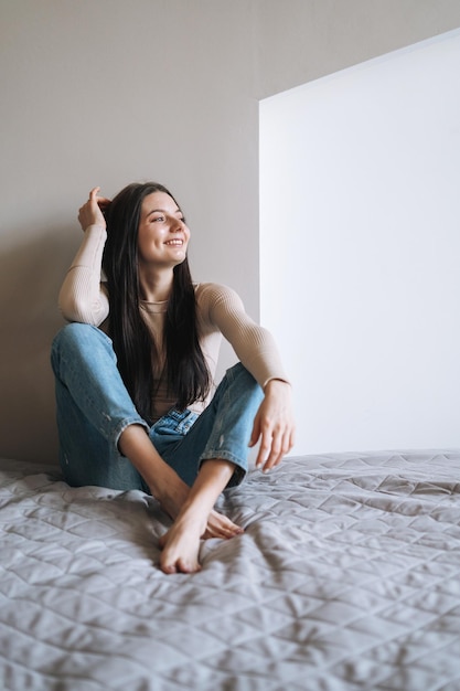 Joven mujer sonriente adolescente con cabello largo oscuro en jeans sentada en la cama en su habitación