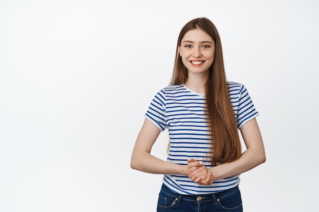 Joven mujer sonriente asistiendo, tomándose de la mano y mirando amigablemente a la cámara, posando contra un fondo blanco.