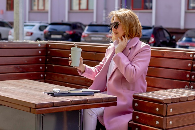 Una joven mujer sonriente bebe café matutino descansando en un banco con el telón de fondo de una ciudad ruidosa