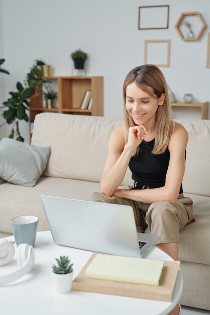 Joven mujer sonriente con cabello largo y rubio sentado en el sofá frente a la mesa con una computadora portátil y mirando la pantalla durante la comunicación en el chat de video