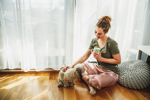 Joven mujer sonriente disfrutando de un café con su perro Shih Tzu en la acogedora mañana en casa.