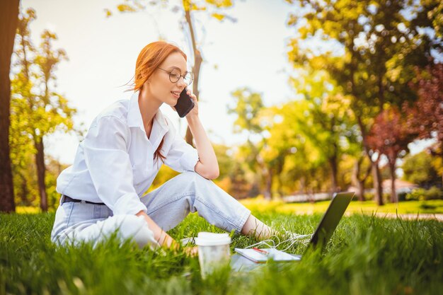 Joven mujer sonriente feliz trabaja con ordenador portátil y teléfono móvil al aire libre mientras está sentado en la hierba