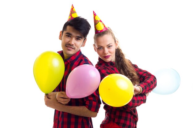 Joven mujer sonriente y hombre agradable con camisas a cuadros rojas con sombreros de celebración que soplan globos