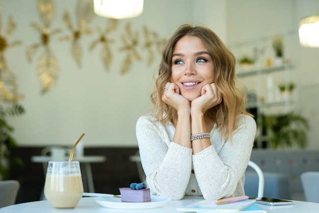 Joven mujer sonriente en ropa casual soñando despierto o simplemente disfrutando del tiempo en la cafetería con un vaso de capuchino y tarta de queso