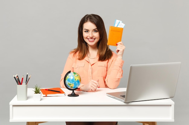 Joven mujer sonriente sosteniendo el globo terráqueo, pasaporte, tarjeta de embarque, billete de trabajo en el escritorio con un ordenador portátil aislado en un fondo gris. Concepto de estilo de vida de carrera empresarial de logro. Simulacros de espacio de copia.