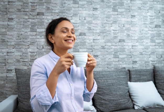 La joven mujer sonriente con una taza de café en sus manos mientras está sentada en el sofá.