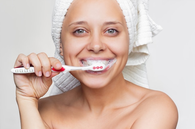 Una joven mujer sonriente con una toalla blanca en la cabeza cepillándose los dientes con un cepillo de dientes