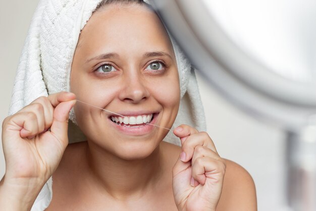 Una joven mujer sonriente con una toalla blanca en la cabeza usando hilo dental sus dientes mirando en el espejo