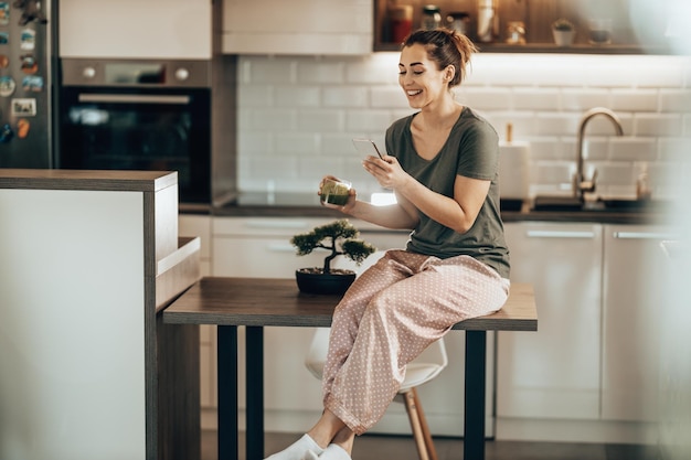 Joven mujer sonriente usando su teléfono inteligente mientras bebe un batido verde por la mañana en casa. bebiendo batido verde mientras usa su teléfono inteligente