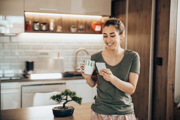 Joven mujer sonriente usando su teléfono inteligente mientras bebe café por la mañana en casa.