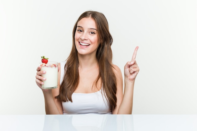 Joven mujer sosteniendo un batido sonriendo alegremente señalando con el dedo lejos