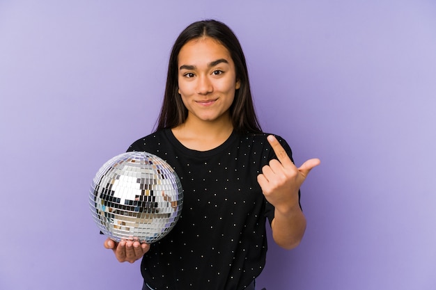 Joven mujer sosteniendo una fiesta de pelota apuntando con el dedo hacia usted como si invitara a acercarse.