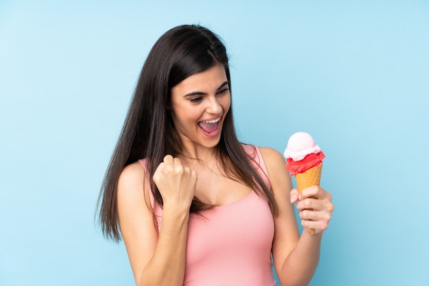 Joven mujer sosteniendo un helado de cucurucho sobre pared azul celebrando una victoria