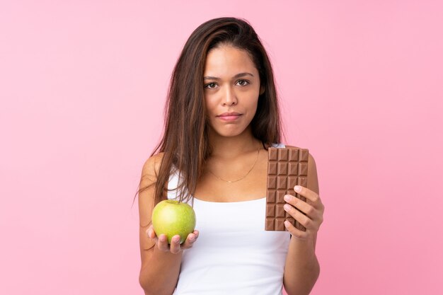 Joven mujer sosteniendo una manzana y chocolate sobre pared aislada