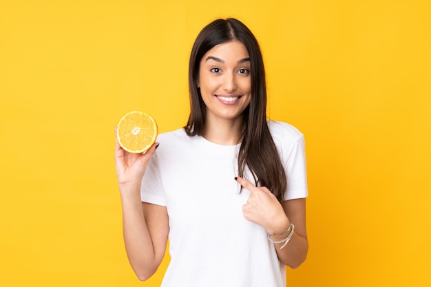 Joven mujer sosteniendo una naranja sobre amarillo con expresión facial sorpresa