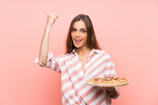 Joven mujer sosteniendo una pizza sobre pared rosa aislado celebrando una victoria