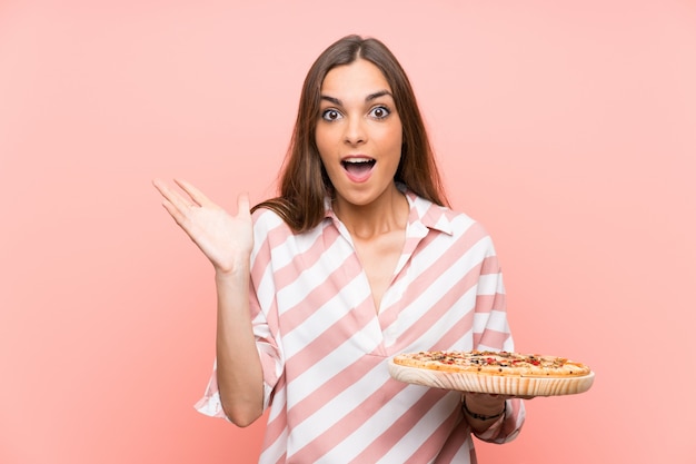 Joven mujer sosteniendo una pizza sobre pared rosa aislado con expresión facial conmocionada