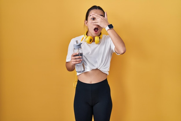 Joven mujer del sur de Asia con ropa deportiva bebiendo agua mirando en shock cubriendo la cara y los ojos con la mano mirando a través de los dedos con expresión avergonzada