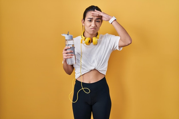 Joven mujer del sur de Asia con ropa deportiva bebiendo agua preocupada y estresada por un problema con la mano en la frente, nerviosa y ansiosa por la crisis