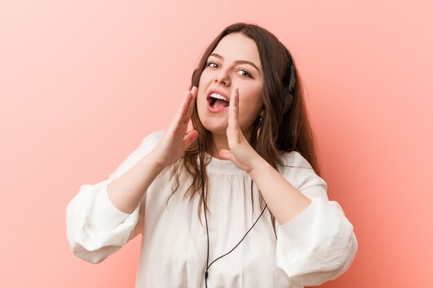 Joven mujer de talla grande con curvas escuchando música con auriculares gritando emocionado al frente.