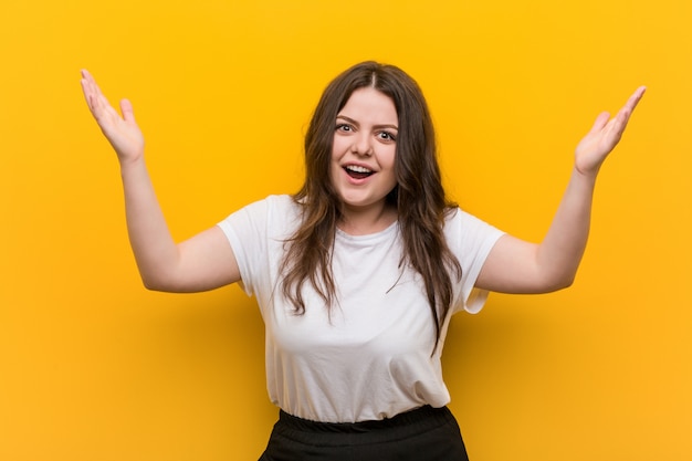 Joven mujer de talla grande con curvas que recibe una agradable sorpresa, emocionada y levantando las manos.