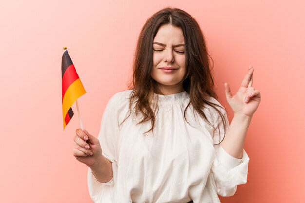 Joven mujer de talla grande con curvas sosteniendo una bandera de Alemania cruzando los dedos para tener suerte