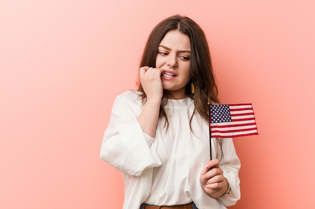 Joven mujer de talla grande con curvas sosteniendo una bandera de los Estados Unidos mordiéndose las uñas, nerviosa y muy ansiosa.