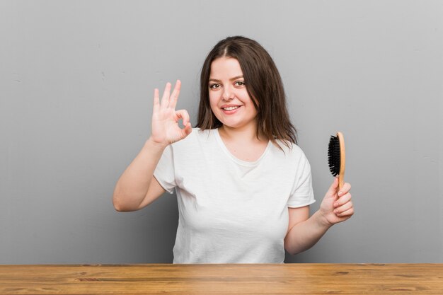 Joven mujer de talla grande con curvas sosteniendo un cepillo para el pelo alegre y confidente que muestra gesto bien.