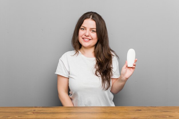 Joven mujer de talla grande con curvas sosteniendo una crema hidratante feliz, sonriente y alegre.