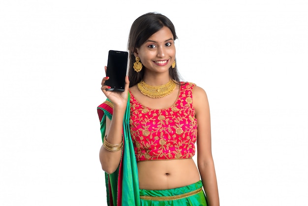 Joven mujer tradicional india usando un teléfono móvil o teléfono inteligente y mostrando el teléfono inteligente de pantalla en blanco sobre blanco