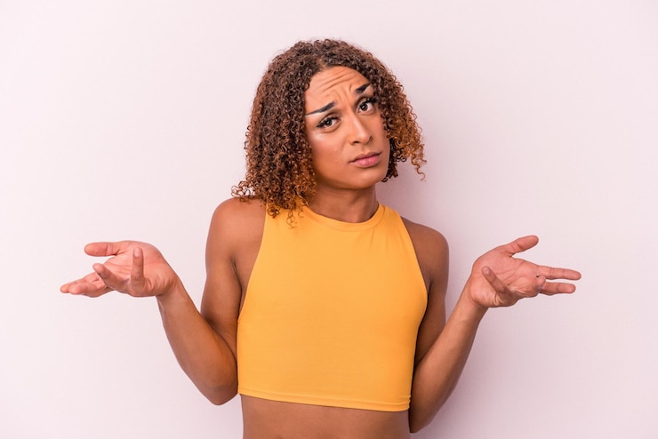Foto joven mujer transexual latina aislada de fondo rosa dudando y encogiéndose de hombros en gesto de cuestionamiento.