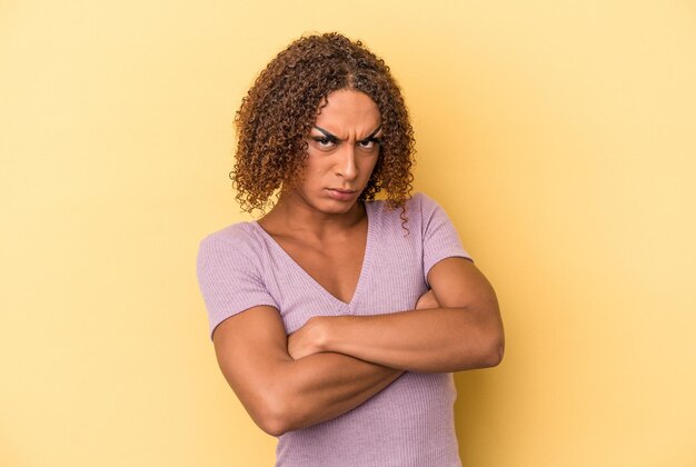 Joven mujer transexual latina aislada sobre fondo amarillo con el ceño fruncido en disgusto, mantiene los brazos cruzados.