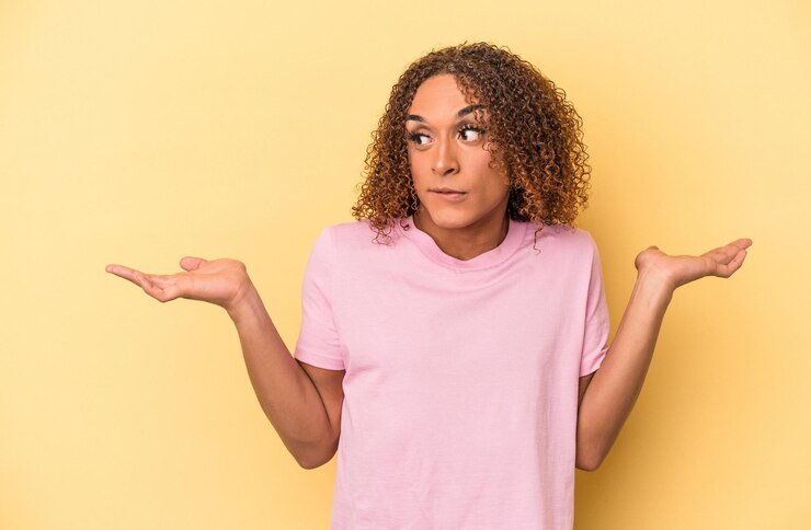 Foto joven mujer transexual latina aislada sobre fondo amarillo confundida y dudosa encogiéndose de hombros para mantener un espacio de copia.