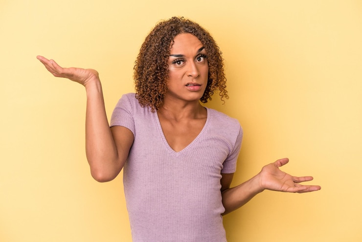 Foto joven mujer transexual latina aislada sobre fondo amarillo confundida y dudosa encogiéndose de hombros para mantener un espacio de copia.