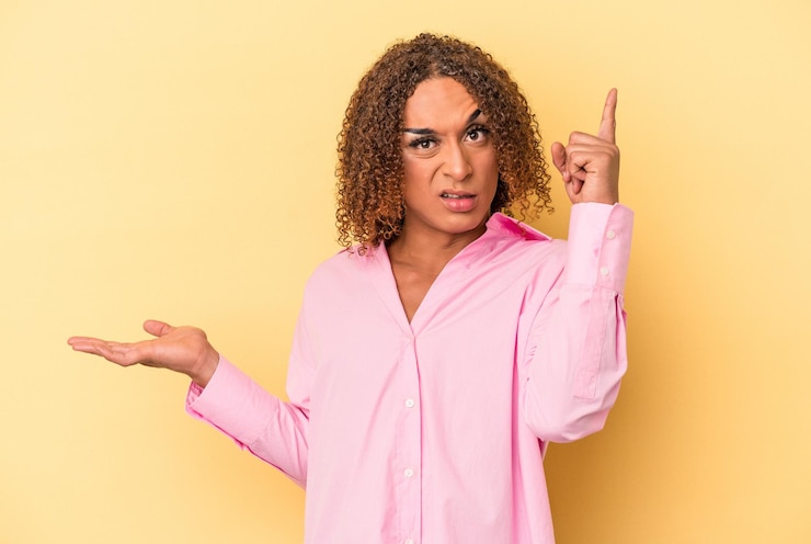 Foto joven mujer transexual latina aislada sobre fondo amarillo sosteniendo y mostrando un producto en la mano.