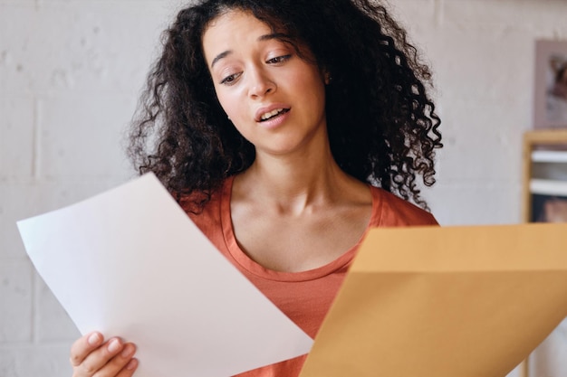 Joven mujer triste con cabello rizado oscuro en camiseta mirando tristemente una carta con resultados de exámenes en las manos en el hogar moderno