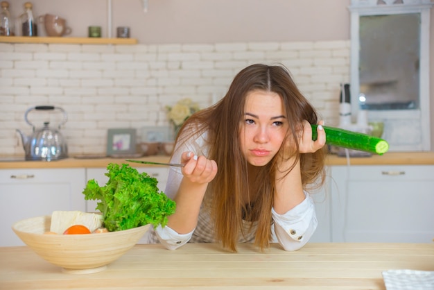 Joven mujer triste comiendo alimentos saludables sentado en el hermoso interior de la cocina. Elección entre comida rápida y comida sana