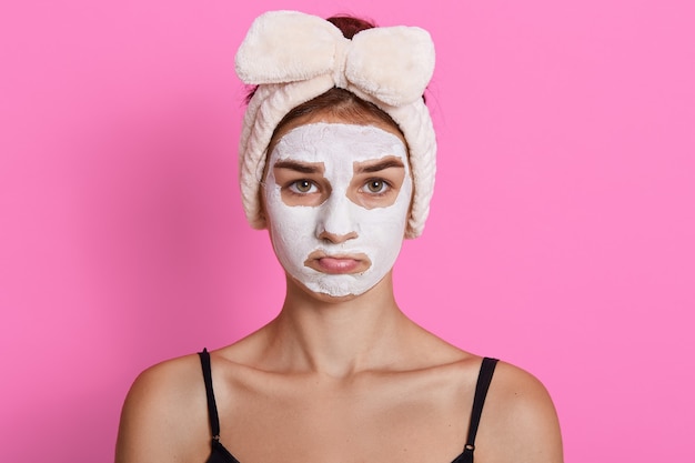 Joven mujer triste con diadema en la cabeza y máscara cosmética en su rostro, posando con labios fruncidos, vistiendo camiseta sin mangas, de pie contra el fondo rosado.