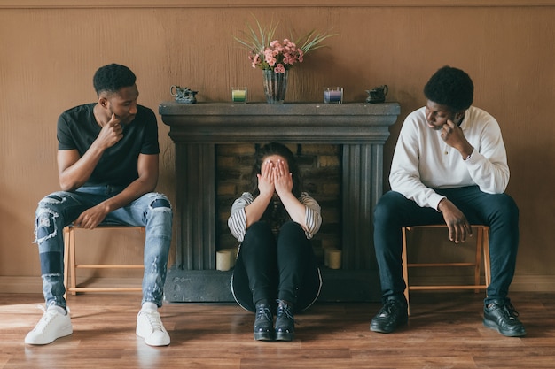 Joven mujer triste sentada en el piso entre dos hombres y cubriendo su rostro llorando con las manos