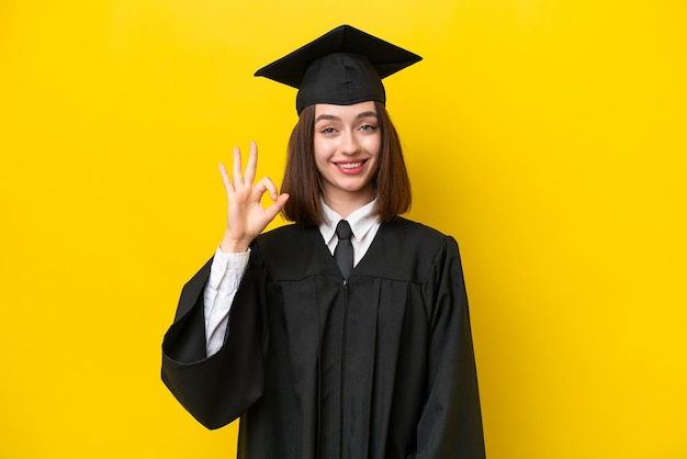 Joven mujer ucraniana graduada de la universidad aislada de fondo amarillo mostrando el signo de ok con los dedos