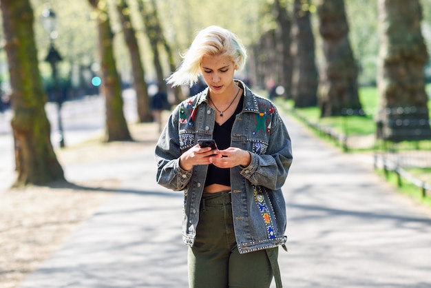 Joven mujer urbana con peinado moderno con smartphone caminando en la calle en un parque urbano.